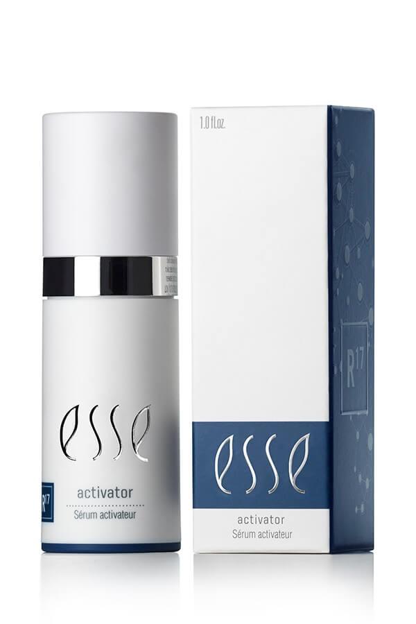 A cylindrical bottle with a metallic cap labeled "Esse Activator Sérum Activateur" is positioned next to its matching box. The box features a blue band at the bottom, showcasing the same text along with a subtle molecular graphic design on the side.