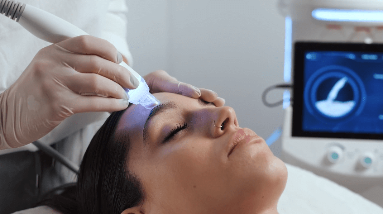 A woman lies with closed eyes as a gloved hand holds the Esse Activator, a modern skincare device emitting blue light, on her forehead. A monitor in the background shows a graphical display. The setting appears to be a professional skincare or cosmetic treatment room.