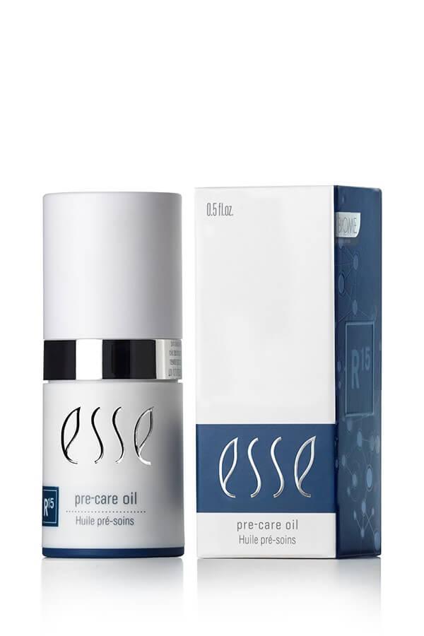 A matte white bottle of Esse Pre-care Oil featuring a metallic silver mirror band in the middle stands next to its matching white and blue box packaging. The box is labeled with "Esse" and "Pre-care Oil," along with a molecular pattern in the background.