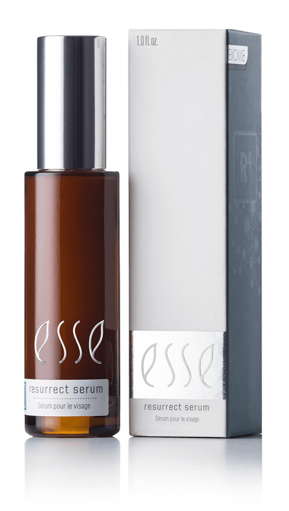 An Esse Resurrect Serum in a brown bottle with a silver cap stands next to its matching white and grey box. Both the bottle's label and the box display the text "Esse Resurrect Serum" along with "Sérum pour le visage," indicating a volume of 1.0 fl oz.