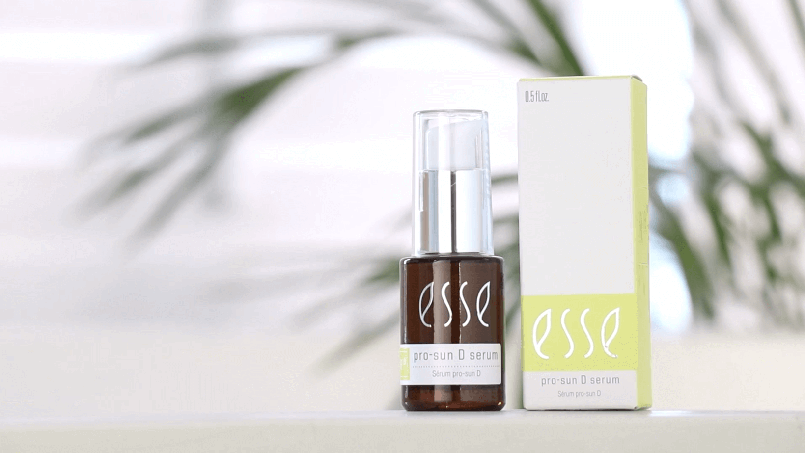A bottle of Esse Pro-Sun D Serum is displayed alongside its box packaging. The brown bottle features white text and a clear cap, whereas the box is mainly white with green accents. In the background, a leafy scene is visible but blurred.