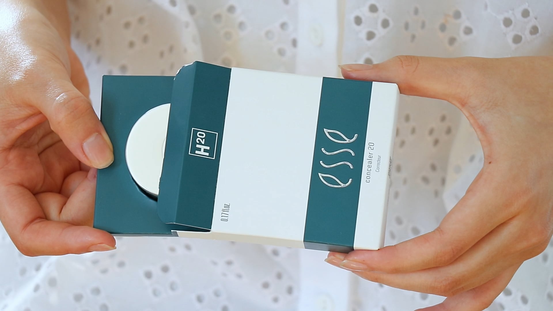 A person holding an open green and white box labeled "H2O" and "Esse Concealer," which appears to contain a circular container. The person is wearing a white, eyelet-patterned shirt with a button front.