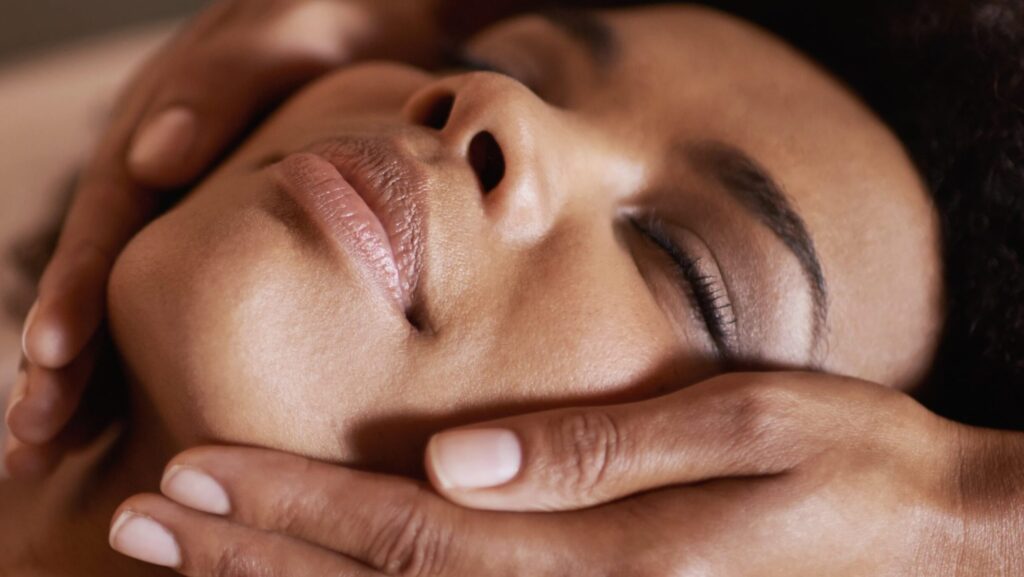 A close-up image of a person receiving a facial massage while using the Esse Dry Skin Trial / Travel Set. The person's eyes are closed, and they appear relaxed. Hands are gently cradling the individual's face, highlighting the comforting nature of the skincare treatment.